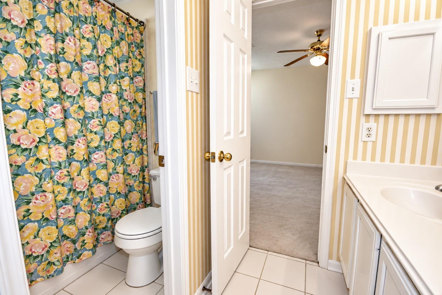 Bathroom with a colorful floral shower curtain, striped walls, and an open doorway to a carpeted room.