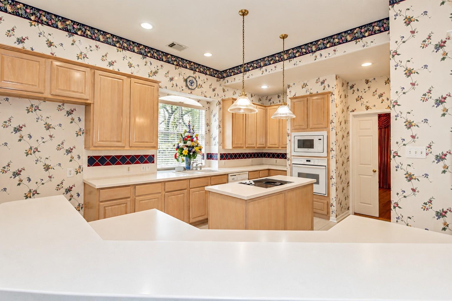 Kitchen with light wood cabinets, floral wallpaper, and a central island with a cooktop.