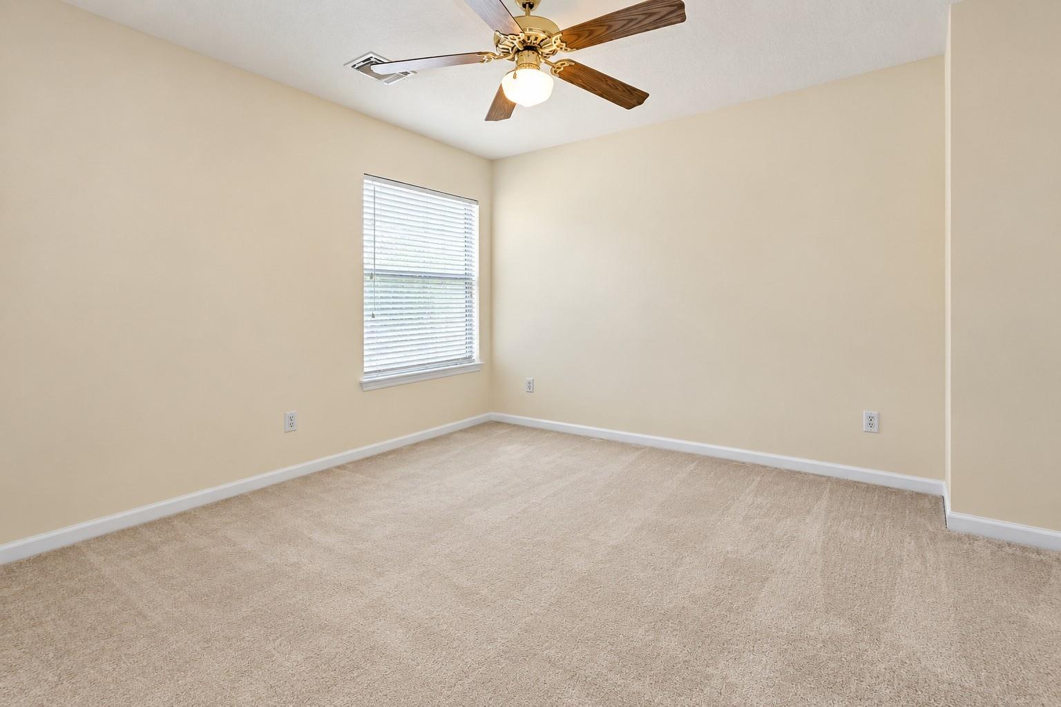 Empty beige-carpeted bedroom with a window and a ceiling fan.