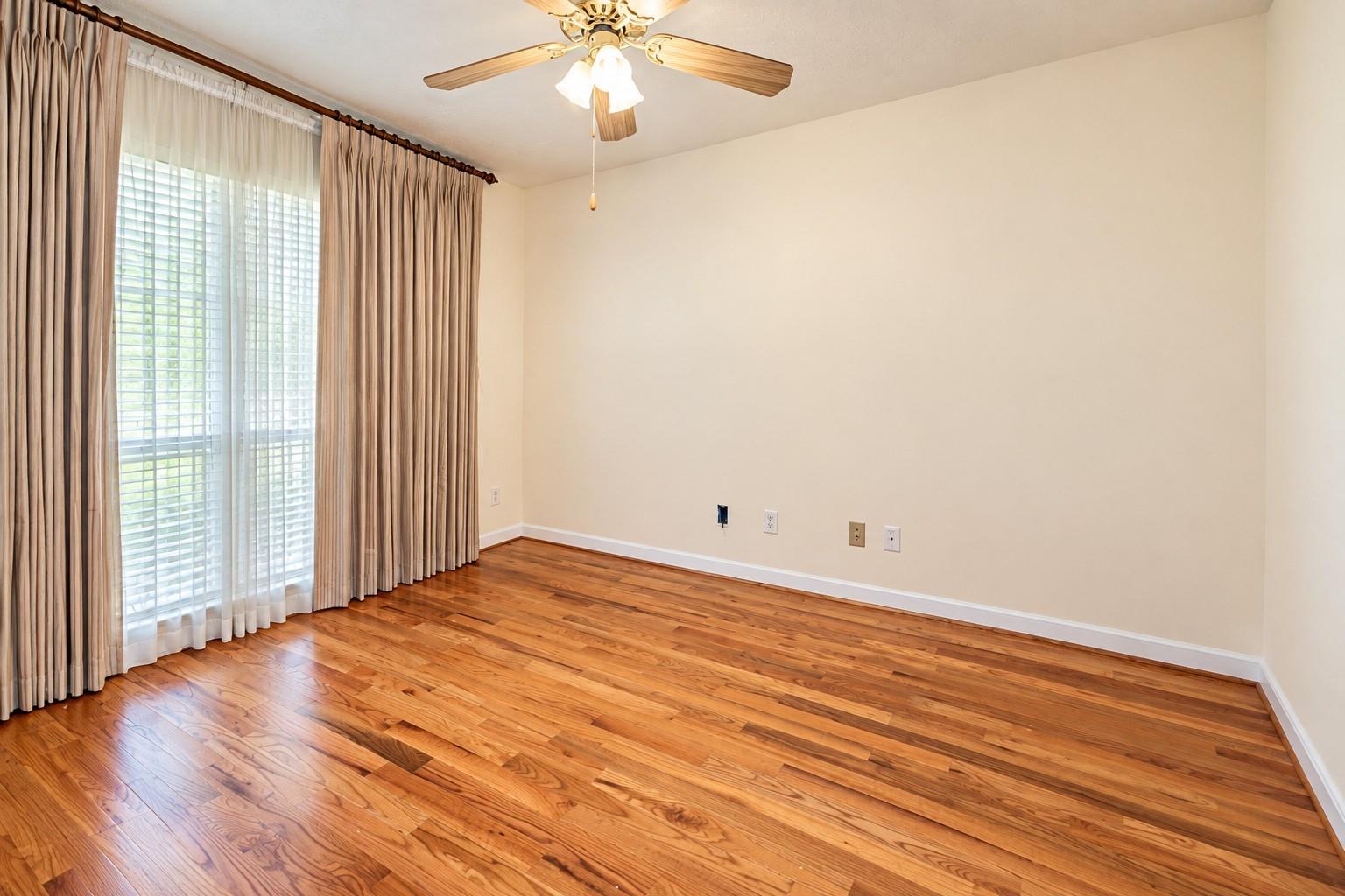 Empty room with hardwood floors, a ceiling fan, and a curtained window.