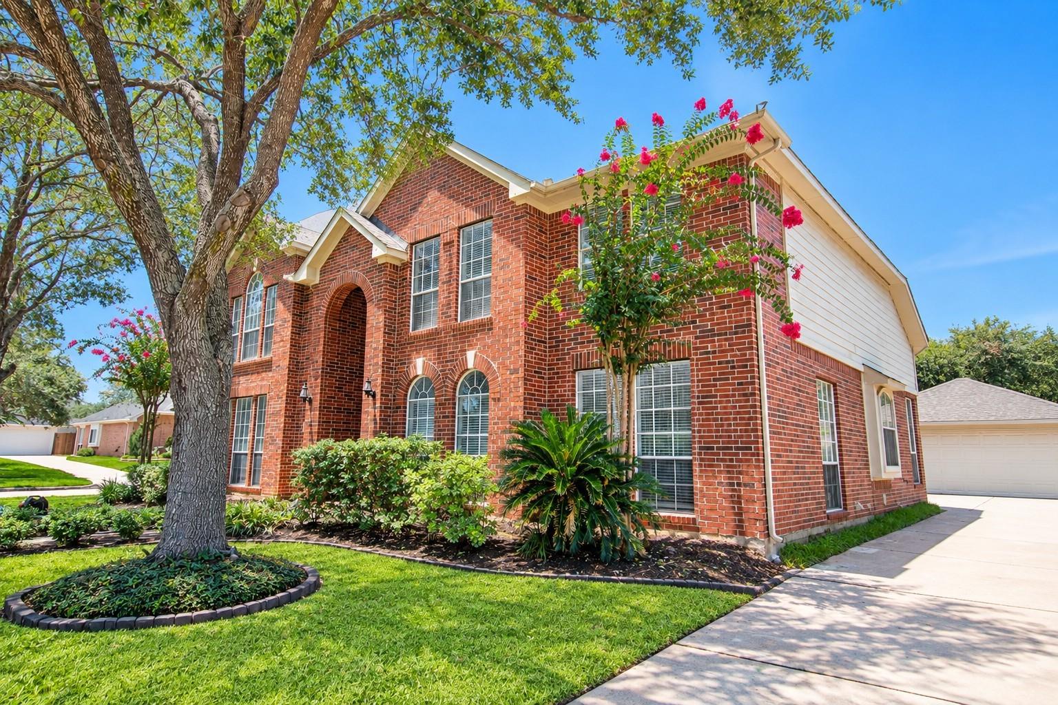 Two-story red brick home with an arched entry, flowering trees, landscaped lawn, and concrete driveway.