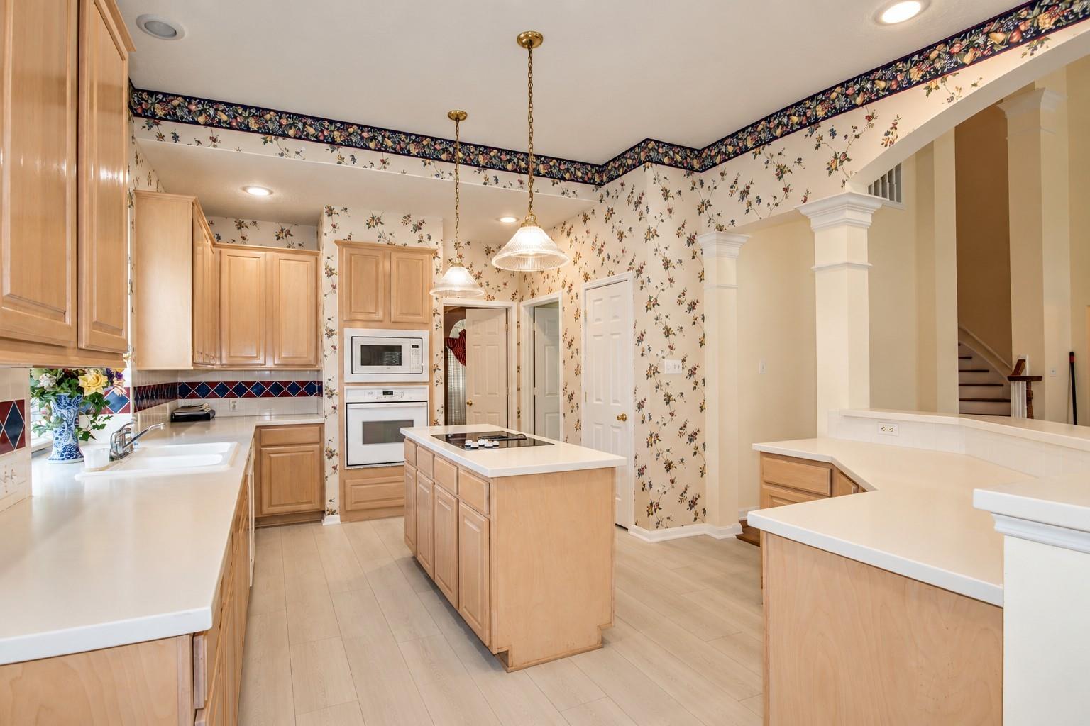 Kitchen with light wood cabinets, a central cooktop island, white counters, and floral wallpaper.