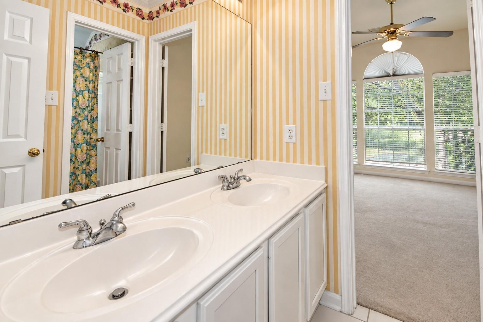 Bathroom with a double-sink vanity, striped wallpaper, and doorway to a bright carpeted room.