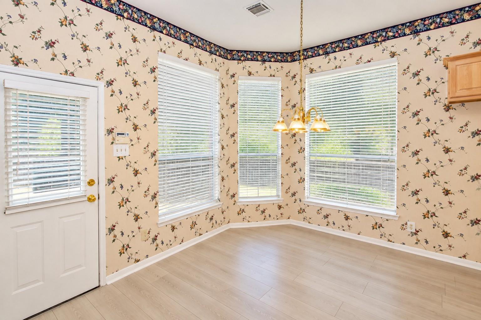 Empty dining area with floral wallpaper, multiple blinded windows, a hanging chandelier, and pale wood flooring.