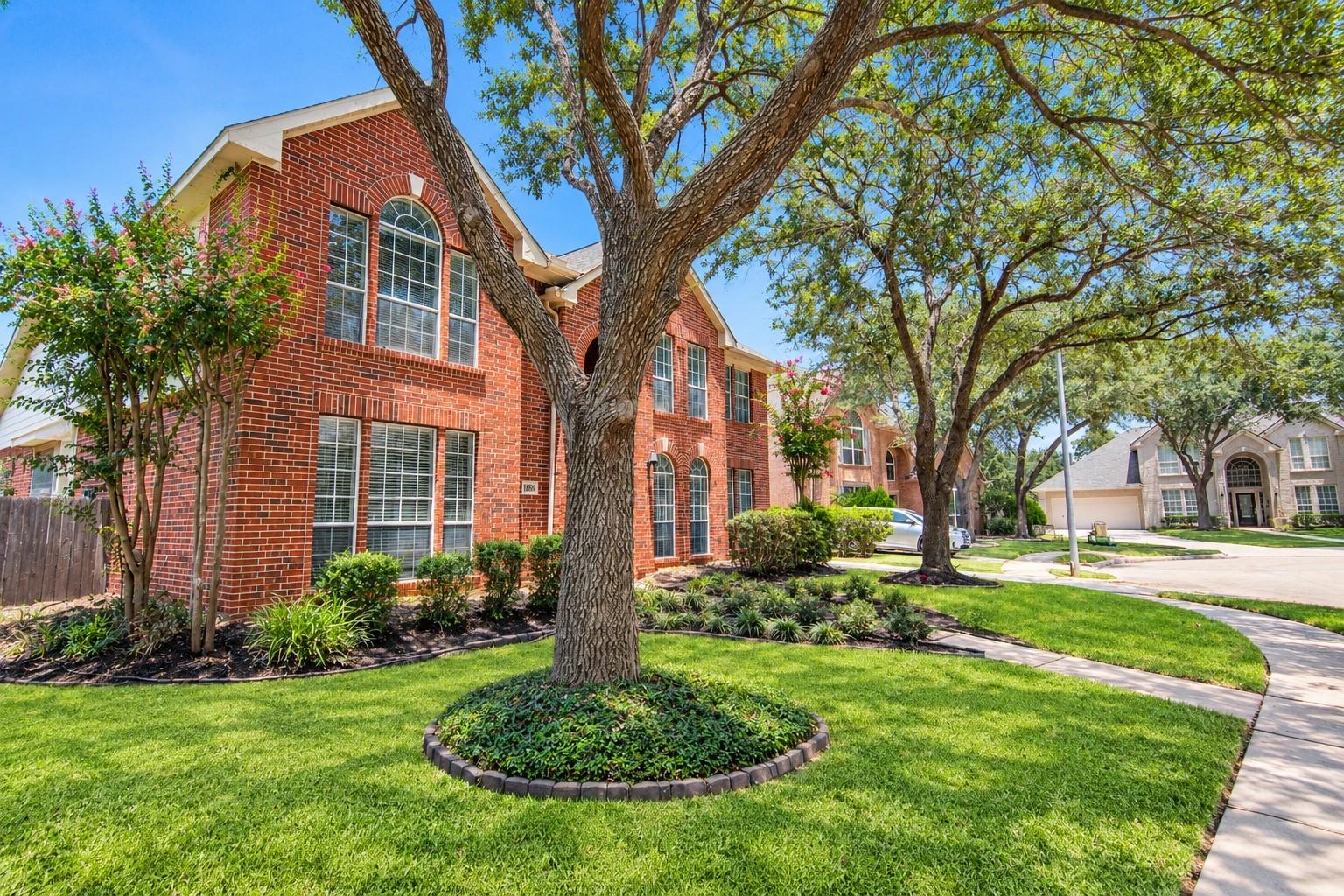 Two-story red brick home with arched windows, mature shade tree, and landscaped lawn.