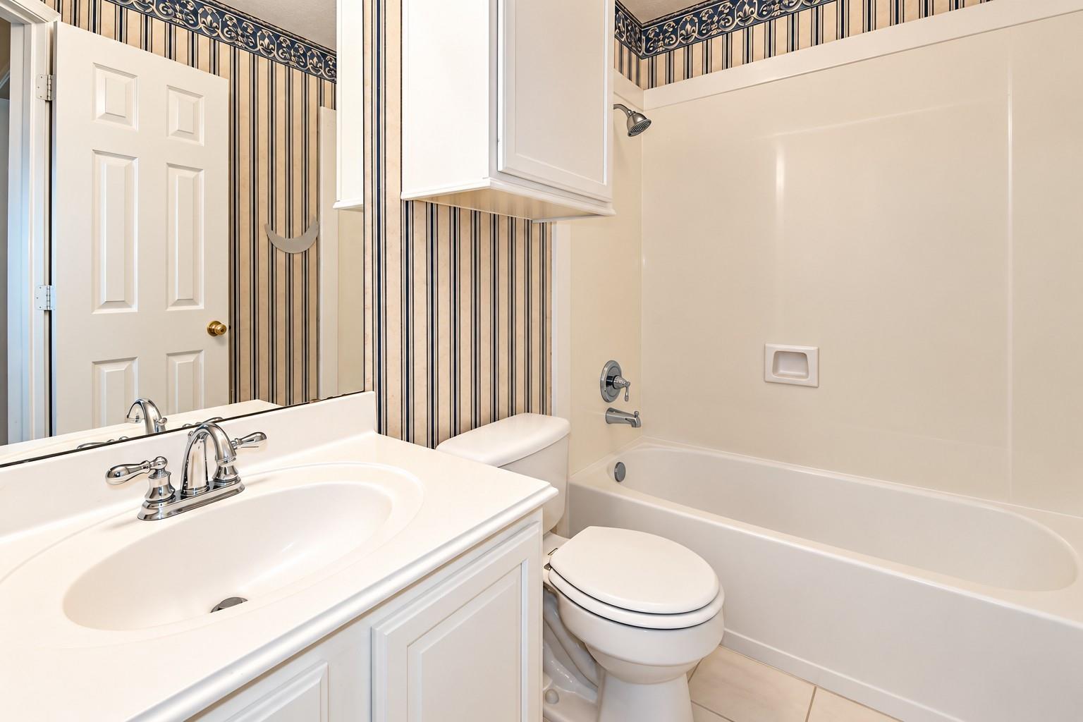 Bathroom with a white vanity, toilet, and tub beside striped wallpaper.