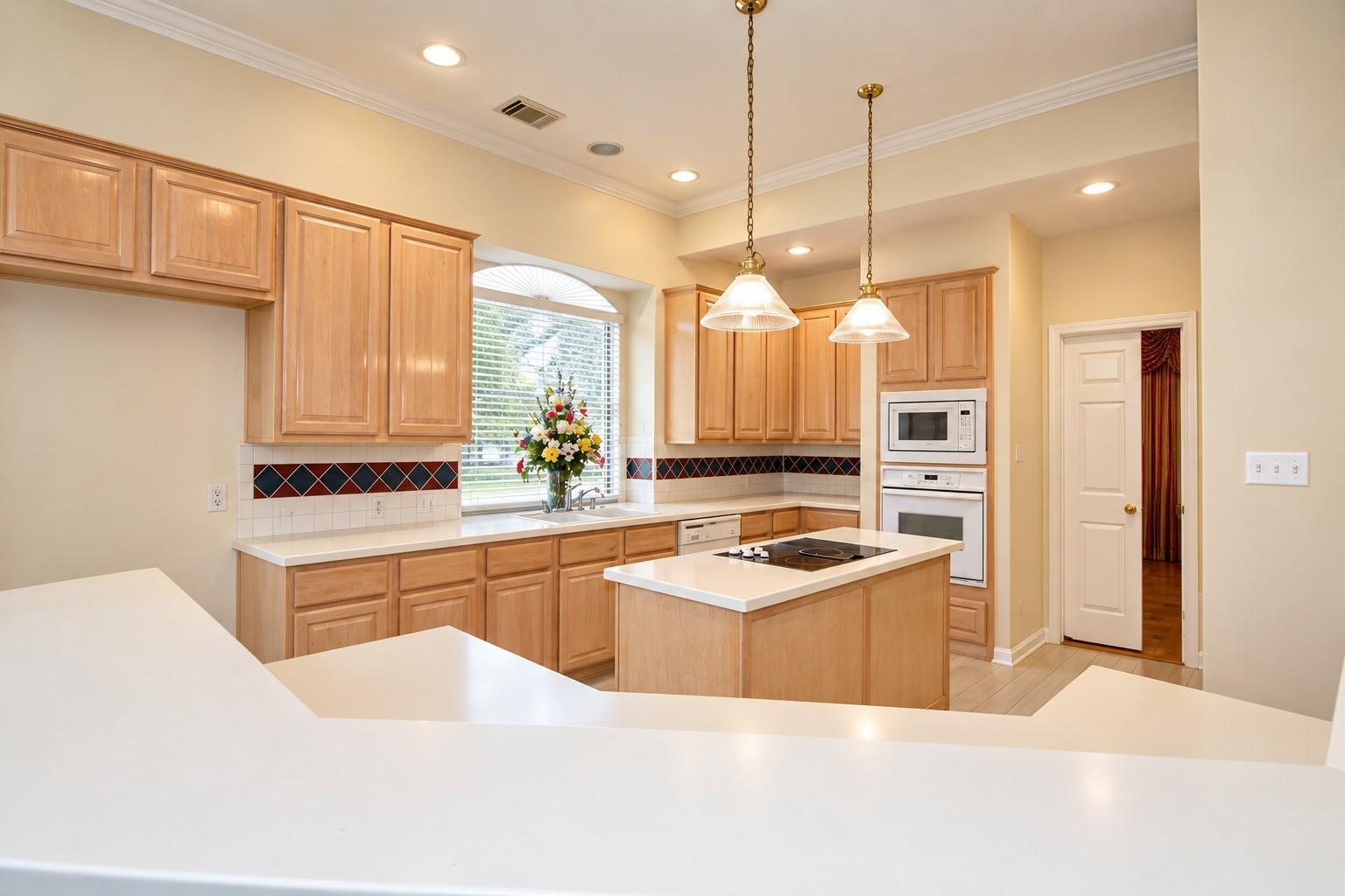 Kitchen with light wood cabinetry, a central cooktop island, and flowers beside the arched window.