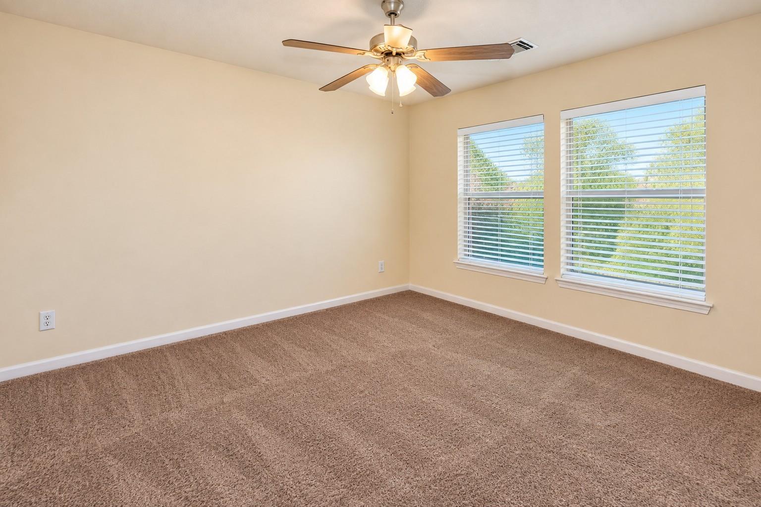 Empty bedroom with brown carpet, two windows with white blinds, and a ceiling fan.