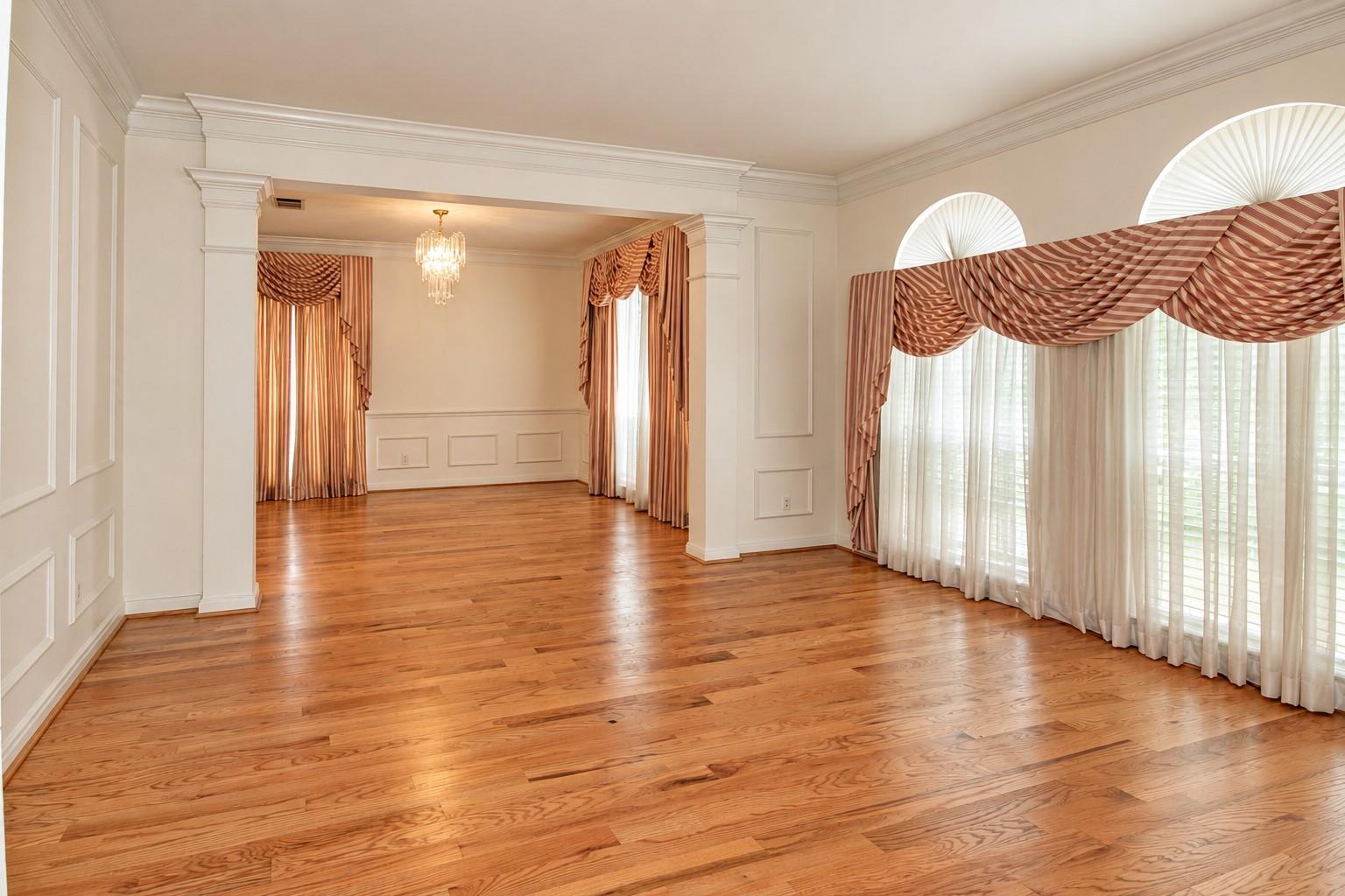 Unfurnished formal room with polished hardwood floors, tall arched windows, and layered curtains.