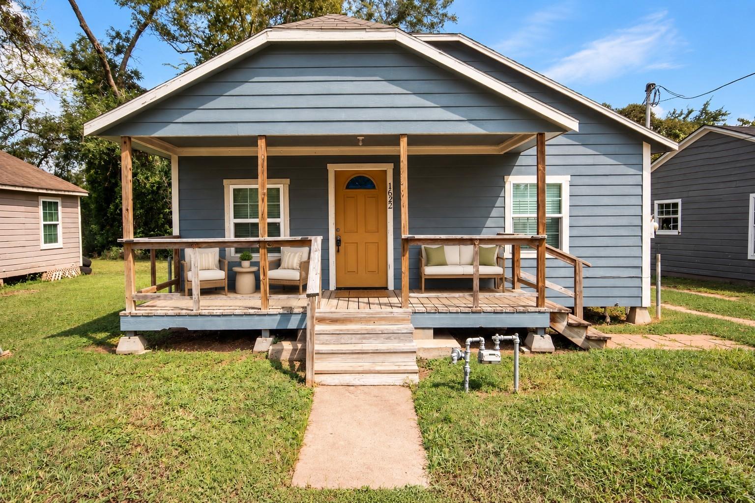 Blue-gray house with a covered porch, wooden steps, outdoor seating, and yellow door marked 1622.