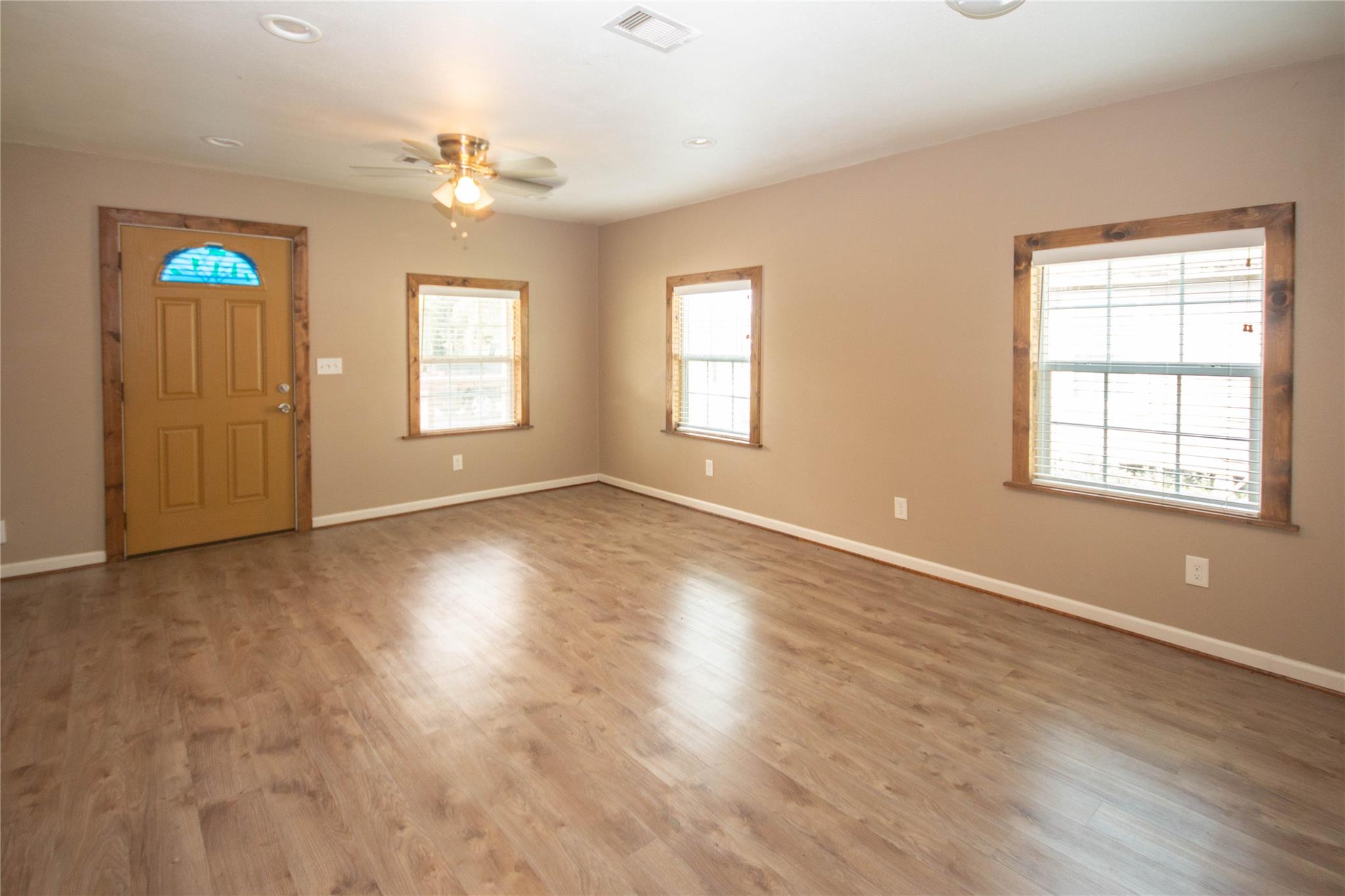 Empty living room with wood-look floors, three wood-trimmed windows, and a front door with stained glass.