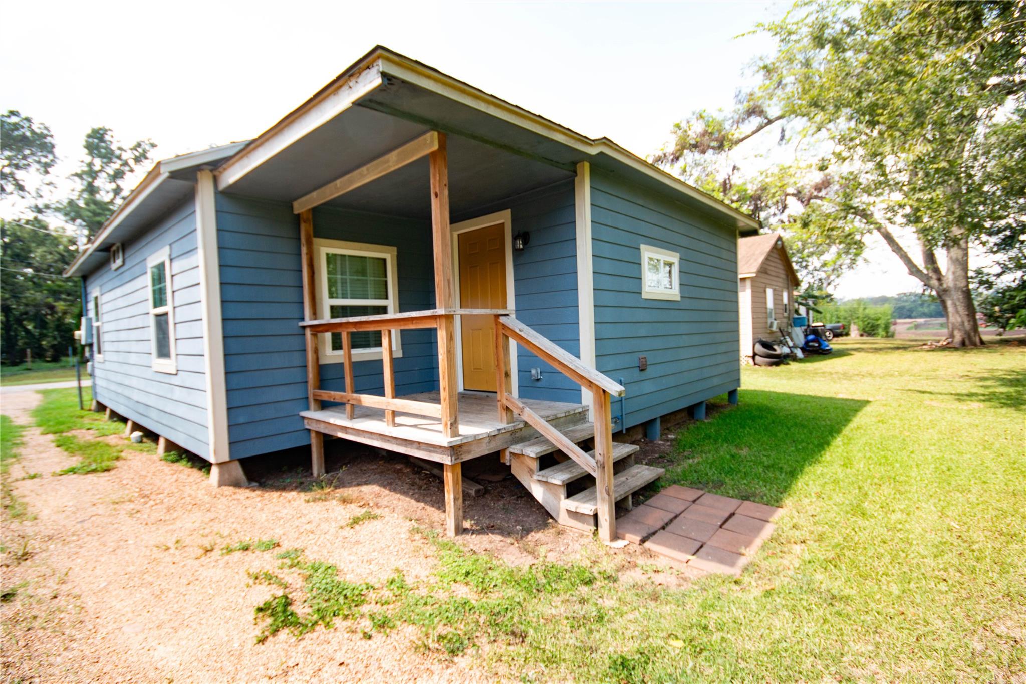 Blue-sided house with a raised wooden porch and front steps beside a sunny grassy yard.