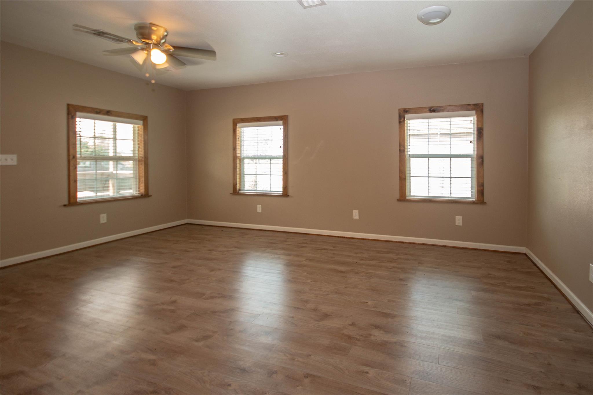 Vacant room with wood-look flooring, three wood-trimmed windows, and a ceiling fan.