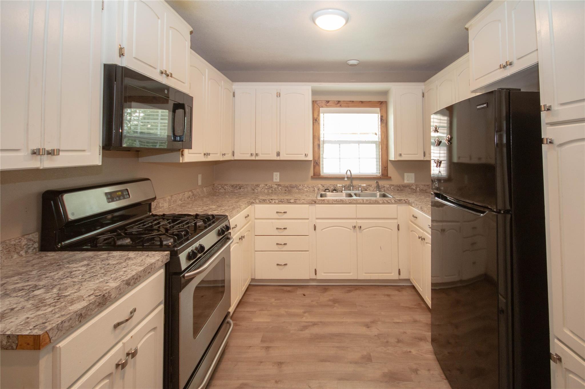 Kitchen with cream cabinets, mottled countertops, black appliances, and a sink beneath a wood-trimmed window.