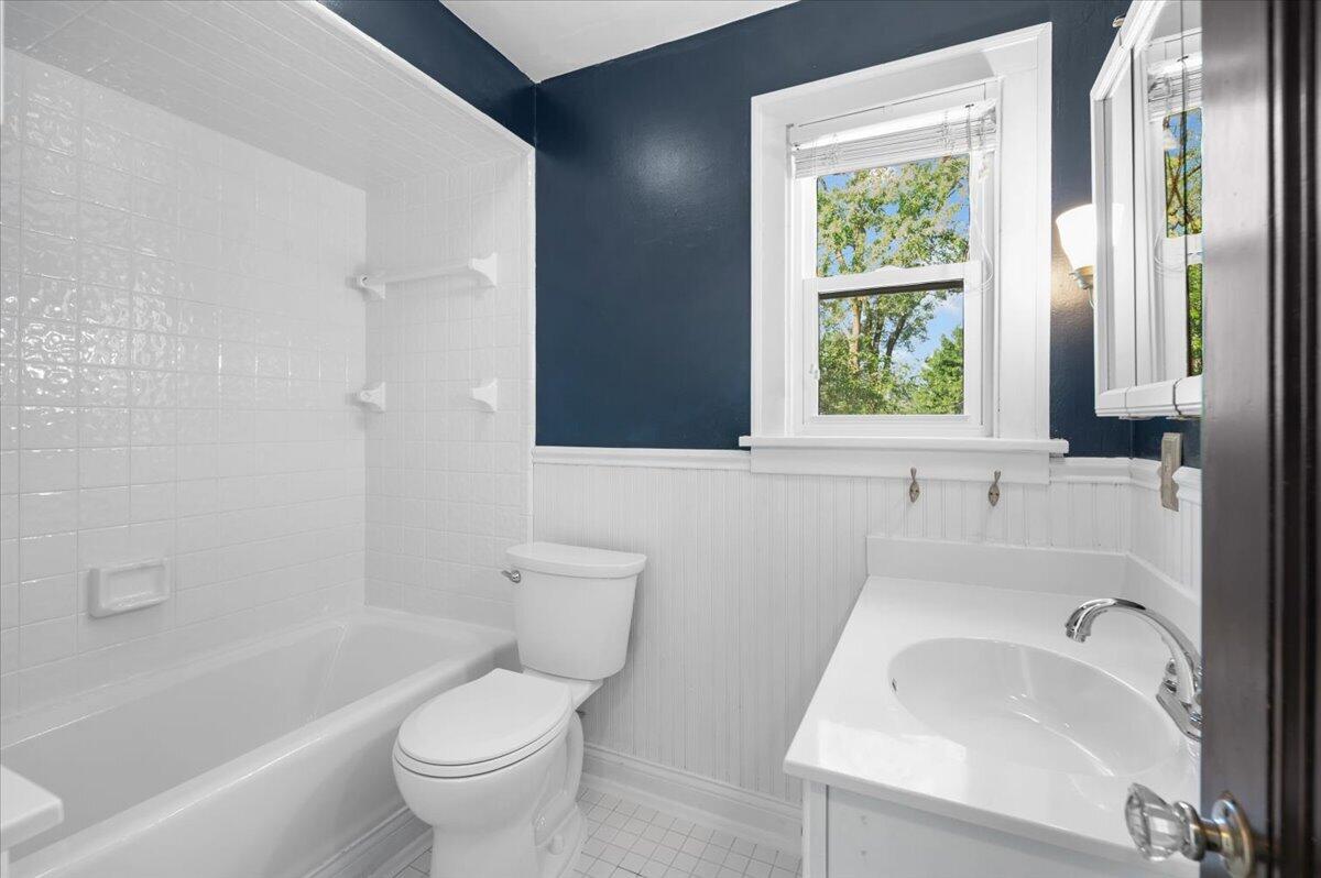 Bathroom with white tiled tub and shower, toilet, and sink beneath a window.