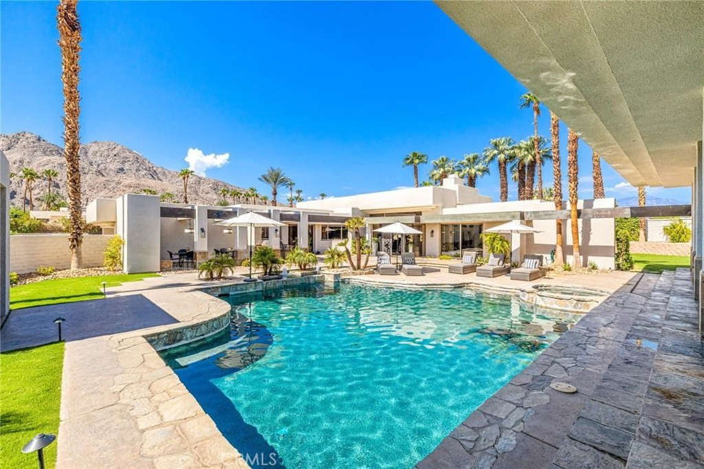 Modern desert home with a turquoise pool, stone patio, palm trees, and mountain backdrop.