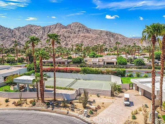 Aerial view of a tennis court and homes with palm trees against desert mountains.