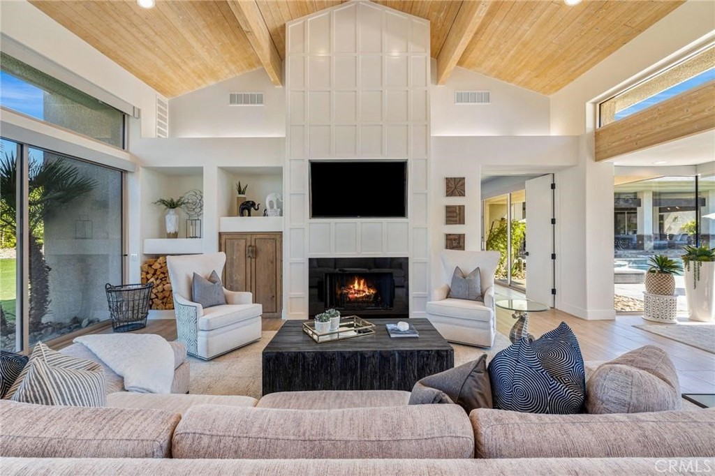 Living room with vaulted wood ceiling, fireplace, built-in television, and large glass doors.