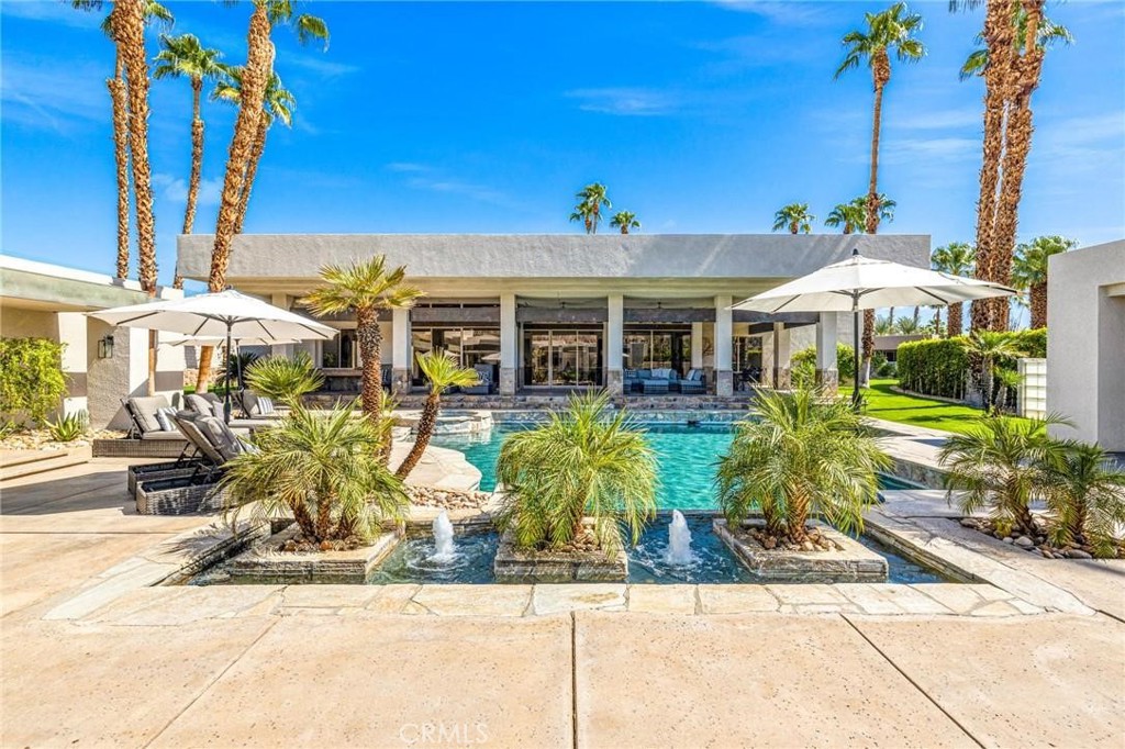 Modern home with a turquoise pool, fountain planters, palm trees, and shaded patio loungers.