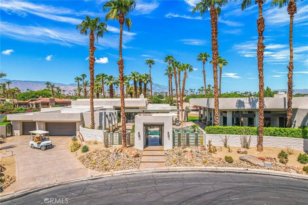 Modern flat-roofed home framed by tall palms, with a gated entry and mountains in the distance.