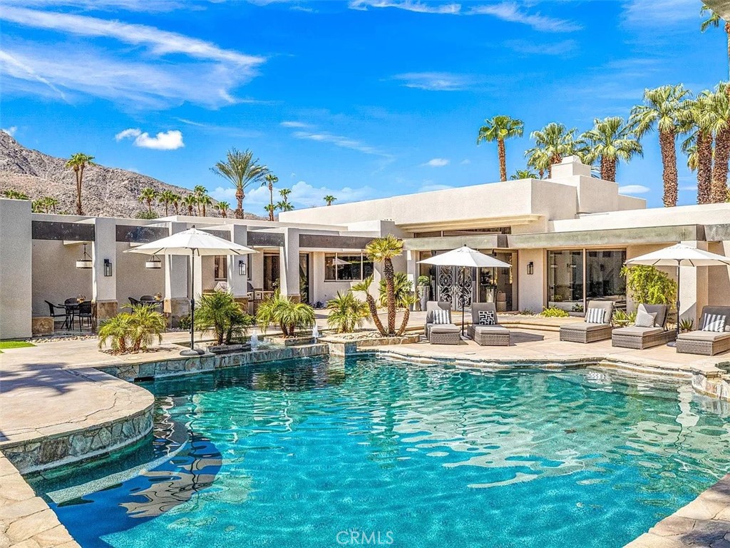 Desert home with a turquoise pool, furnished patio, palm trees, and rocky mountain backdrop.