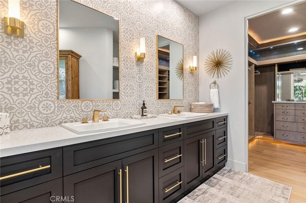Bathroom with a dark double vanity, patterned wallpaper, brass fixtures, and an adjoining walk-in closet.