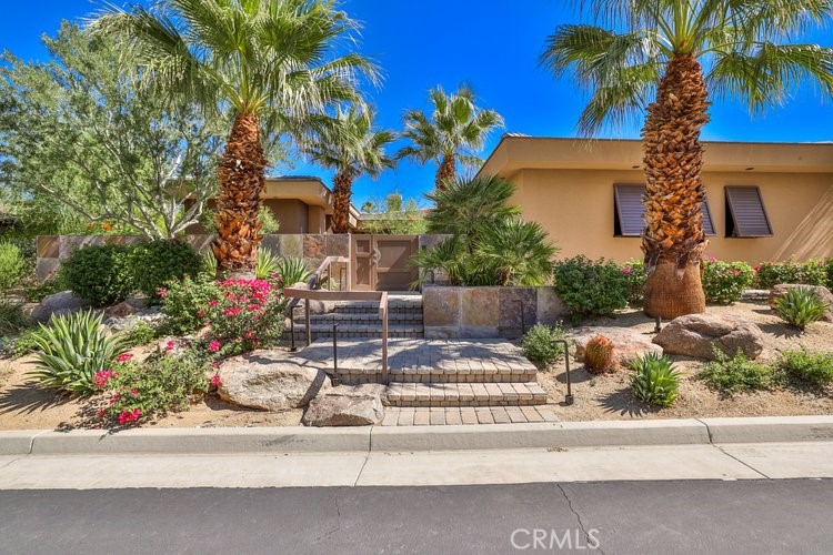 Tan home entrance with stone steps, palm trees, flowering shrubs, and desert landscaping.