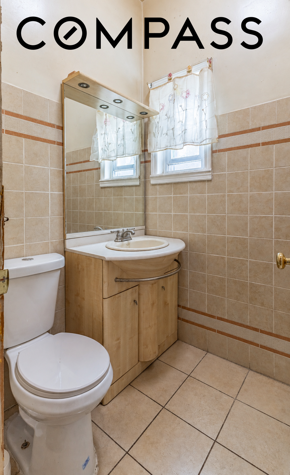 Bathroom with beige tile, wood vanity, large mirror, toilet, and two curtained windows.