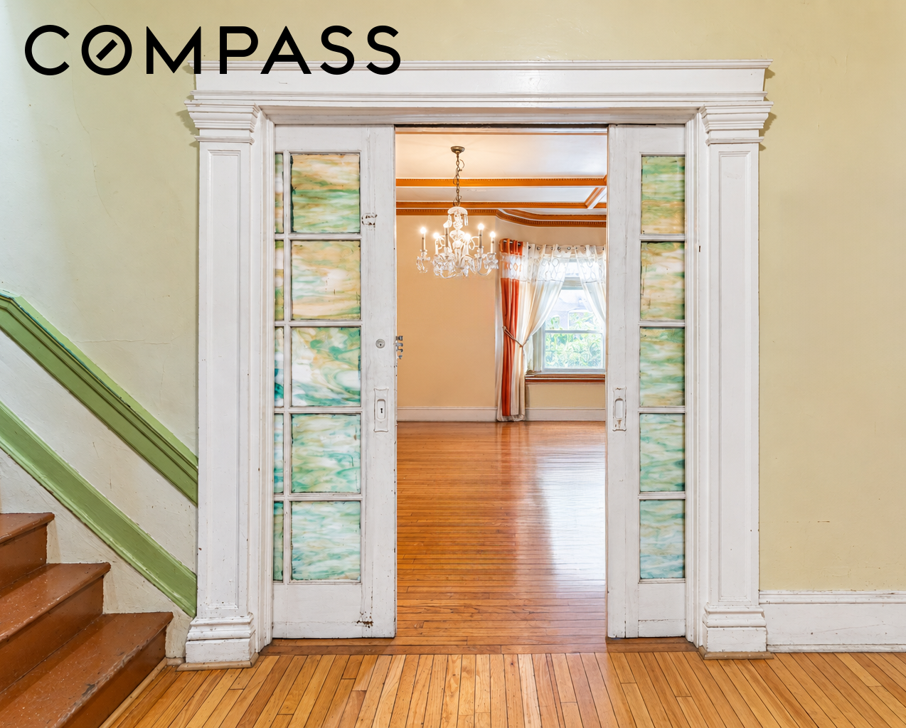Ornate glass-paneled doorway opens to a hardwood-floored room with a chandelier and curtained window.