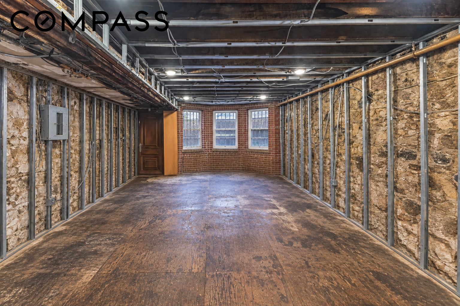 Unfinished room with exposed framing and insulation, worn wood floor, and three windows in a brick wall.