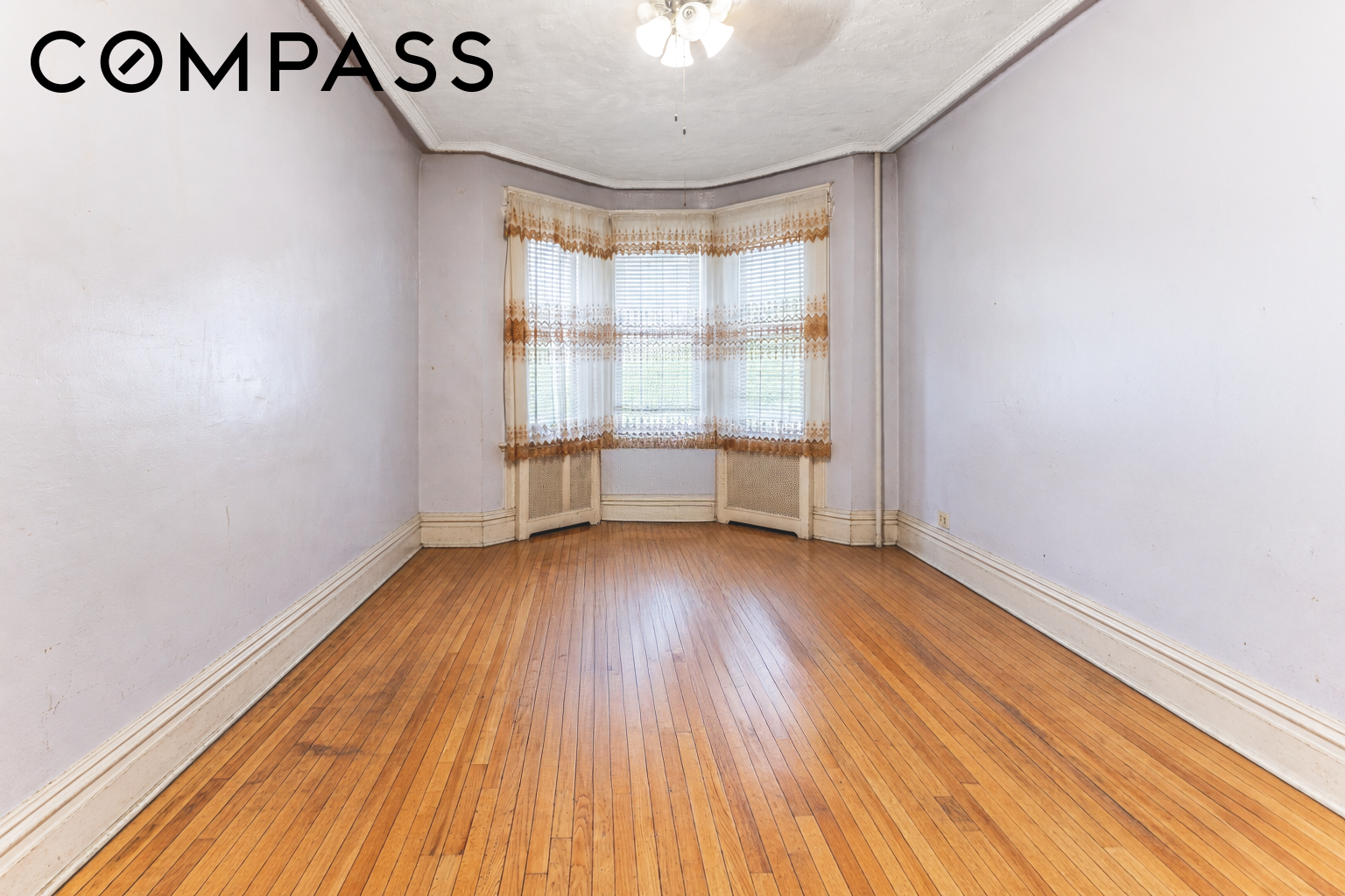Empty room with a bay window, lace curtains, and polished hardwood floors.