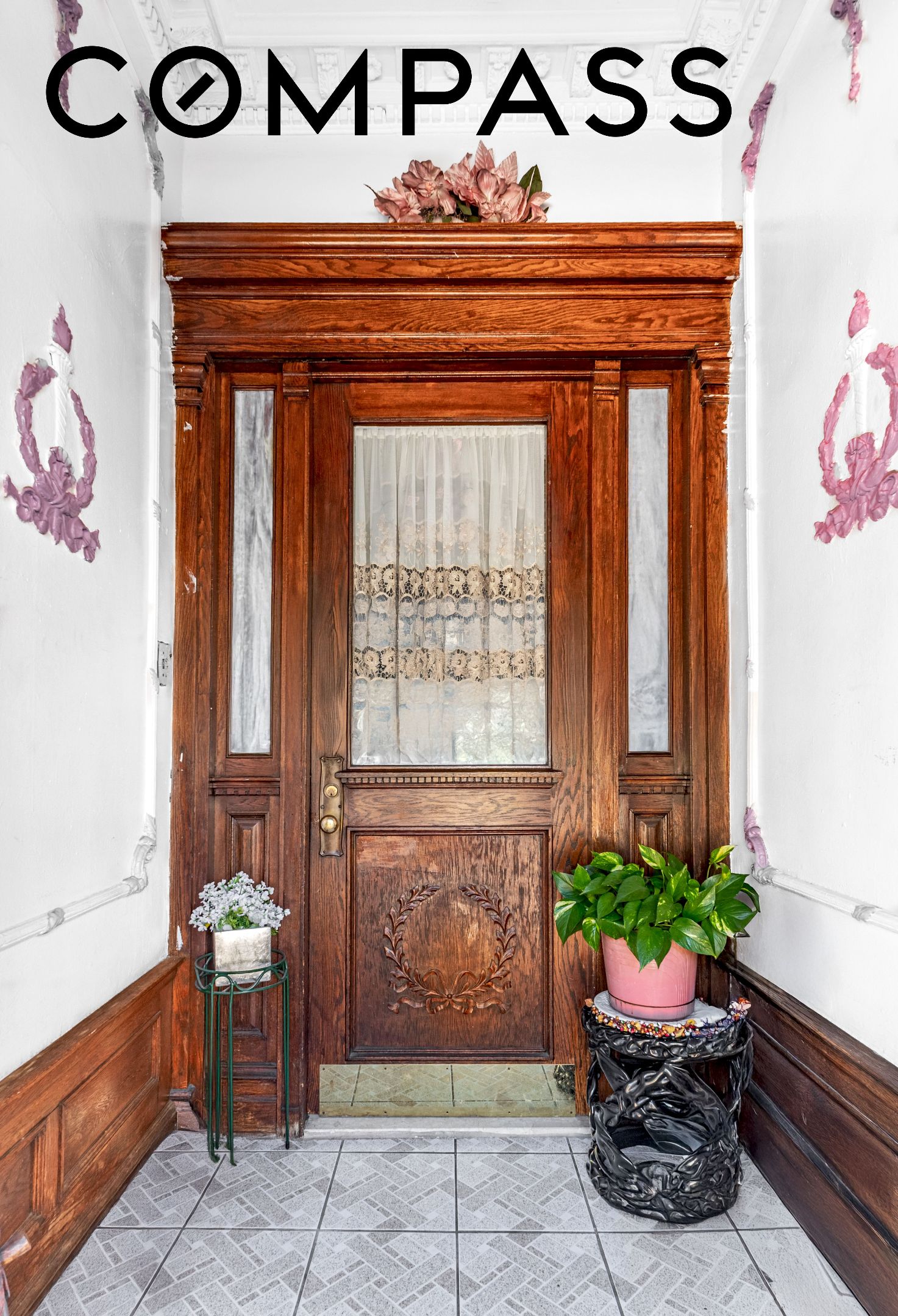 Entryway with an ornate carved wooden door, glass sidelights, potted plants, and patterned tile floor.