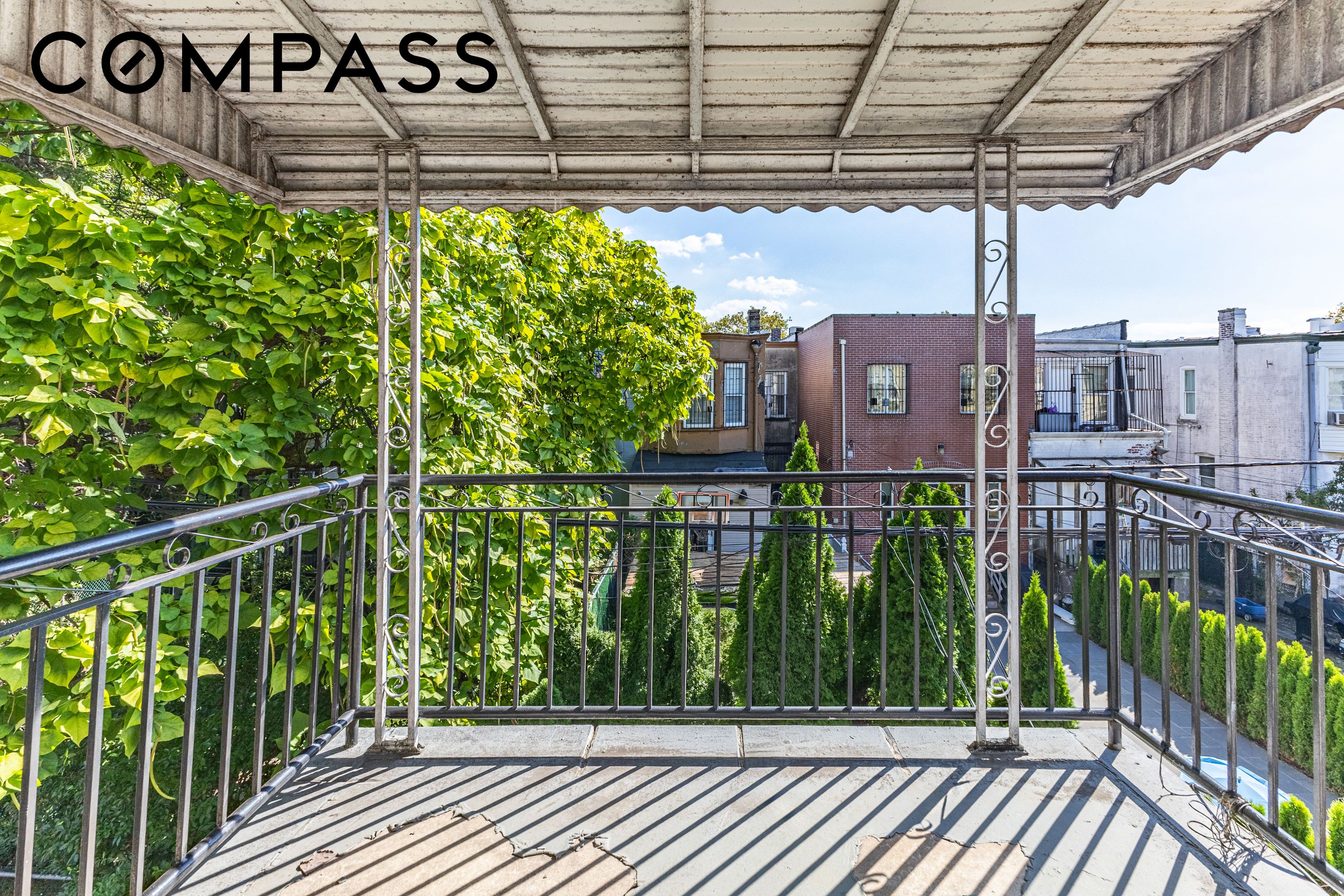 Covered balcony with ornate metal railings, leafy greenery, and neighboring rooftops.