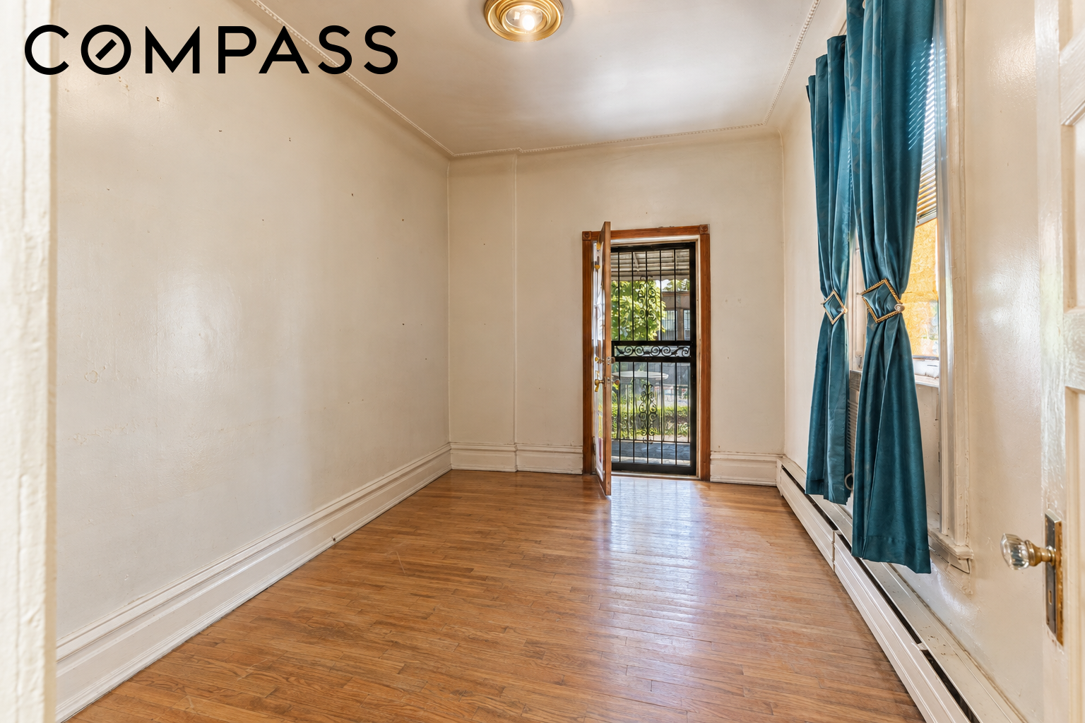 Empty room with hardwood floors, teal curtains, and an open doorway with a metal security gate.