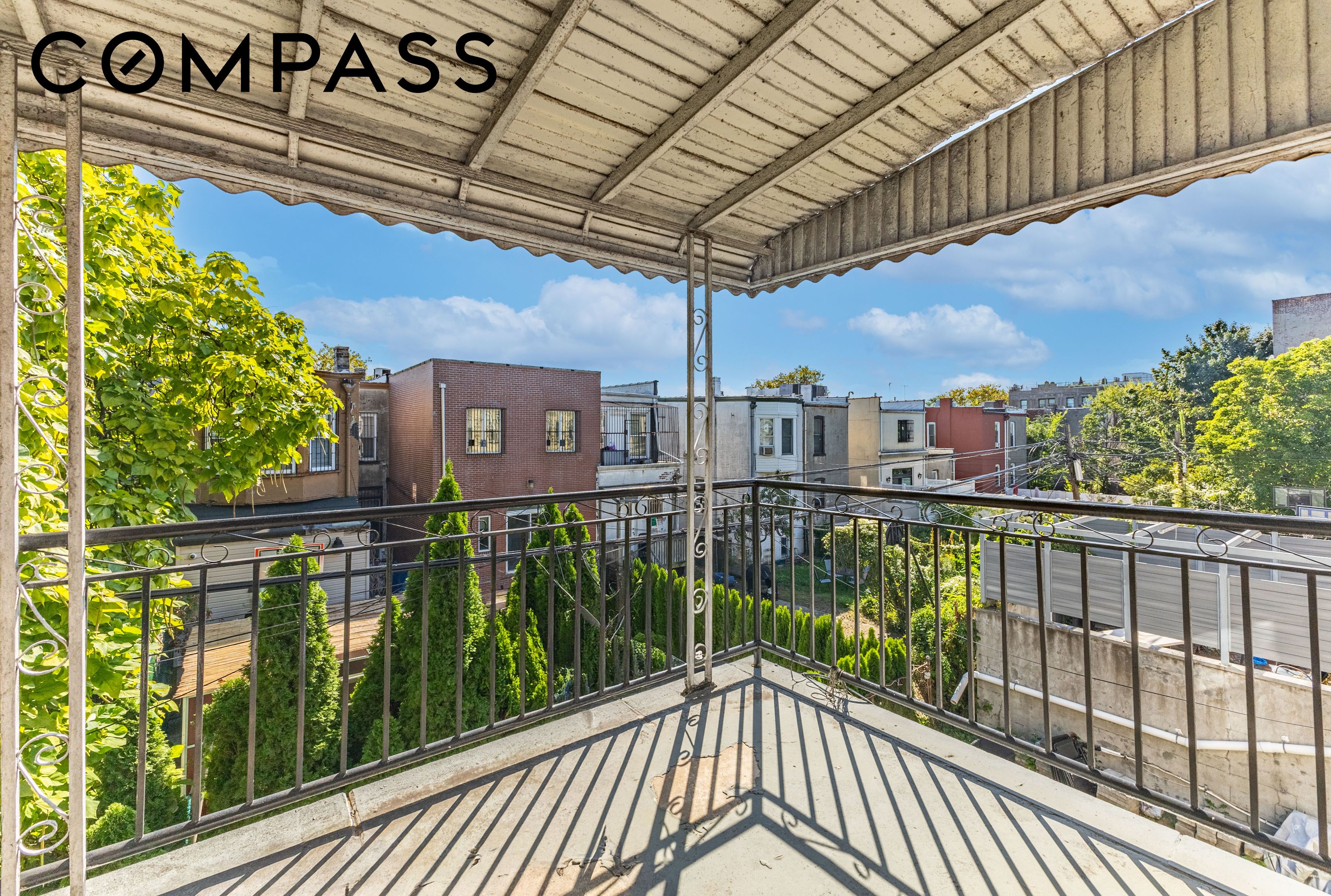 Covered balcony overlooking leafy yards and neighboring rooftops, with a metal railing and striped shadows.