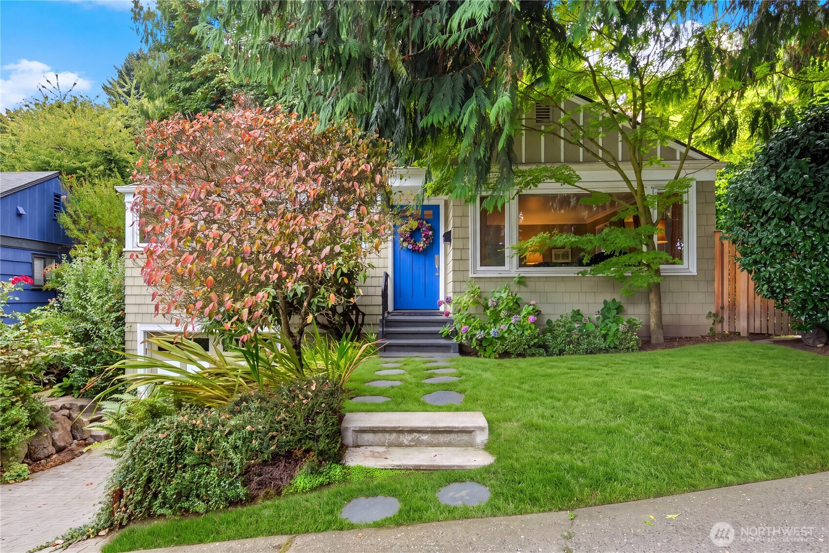 Shingle-sided home with a bright blue front door, leafy landscaping, stepping stones, and a green lawn.