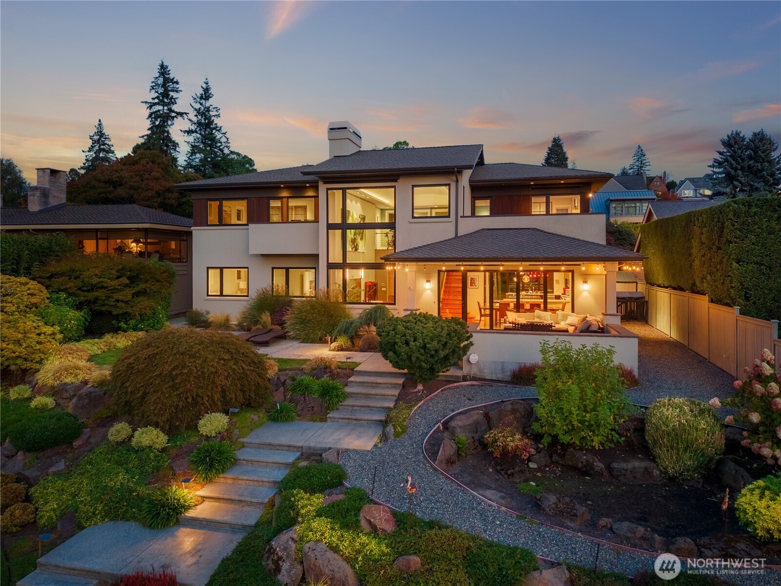 Modern multi-story home with a covered patio and landscaped terraced yard at dusk.