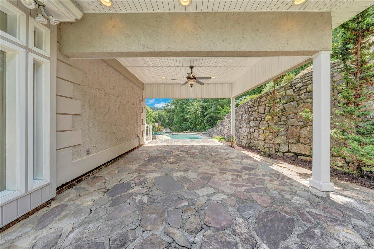 Covered flagstone patio with a ceiling fan, stone retaining wall, and swimming pool beyond.