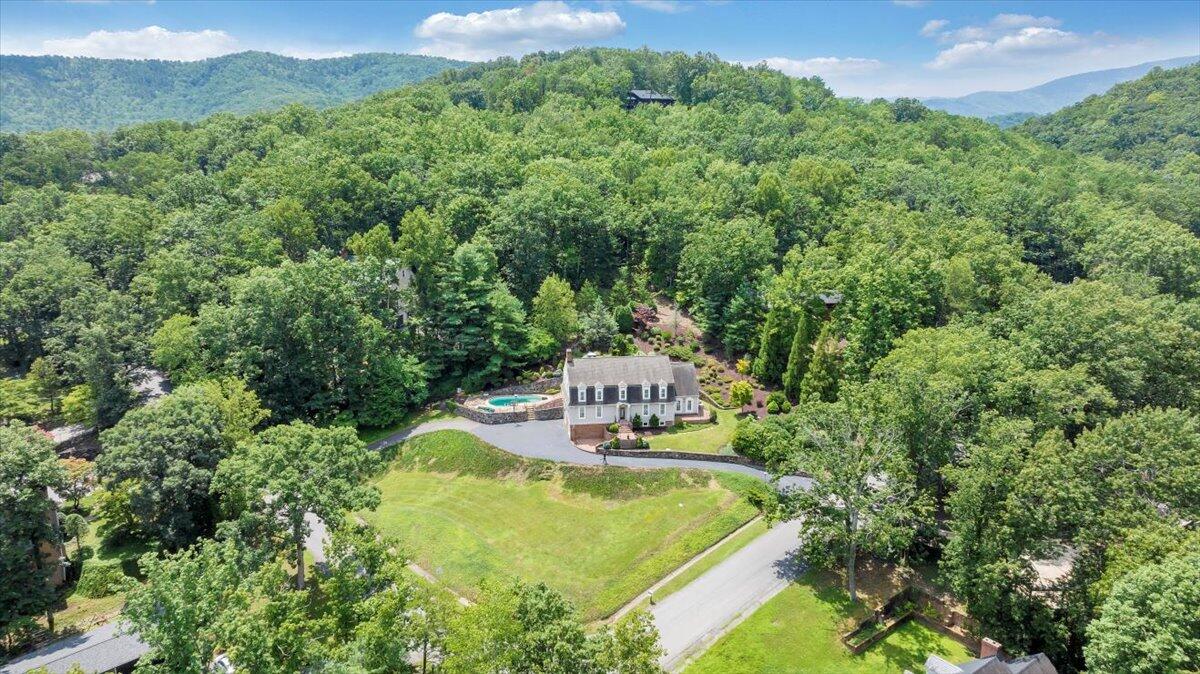 Aerial view of a house with a swimming pool, broad lawn, and forested mountain backdrop.
