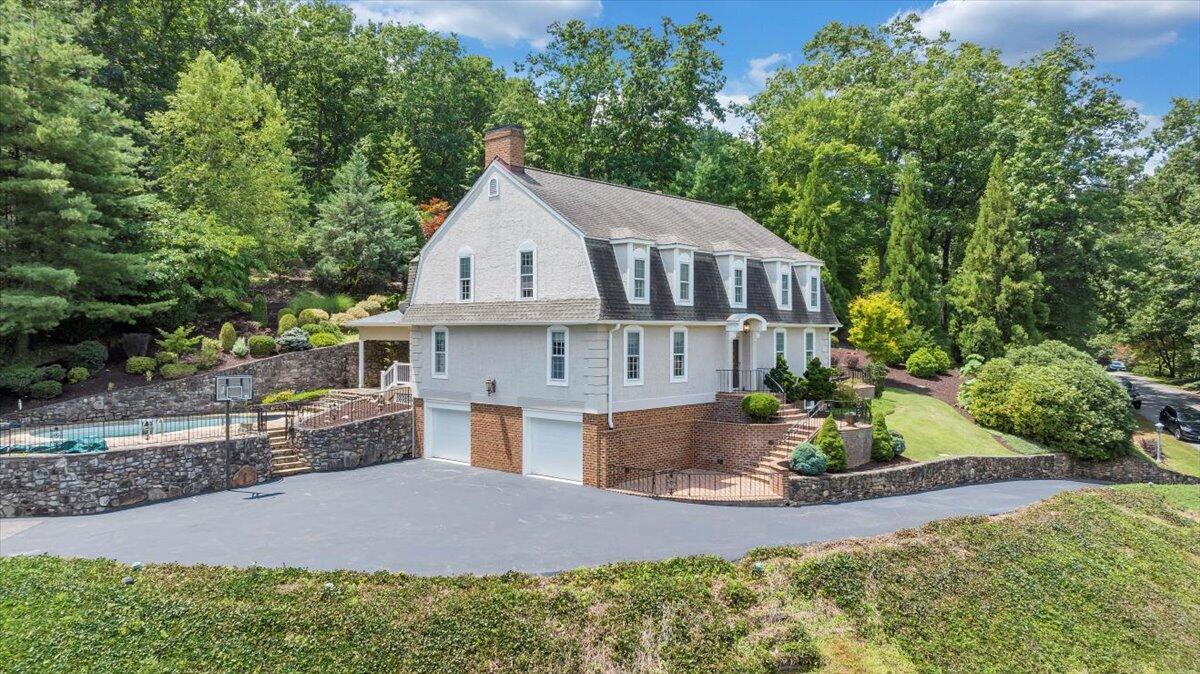 White-and-brick home with dormer windows, a pool, and stone retaining walls amid mature trees.
