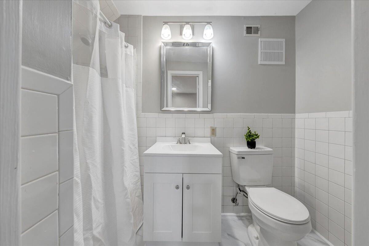 Bathroom with white vanity, tiled walls, shower curtain, and toilet topped with a small plant.