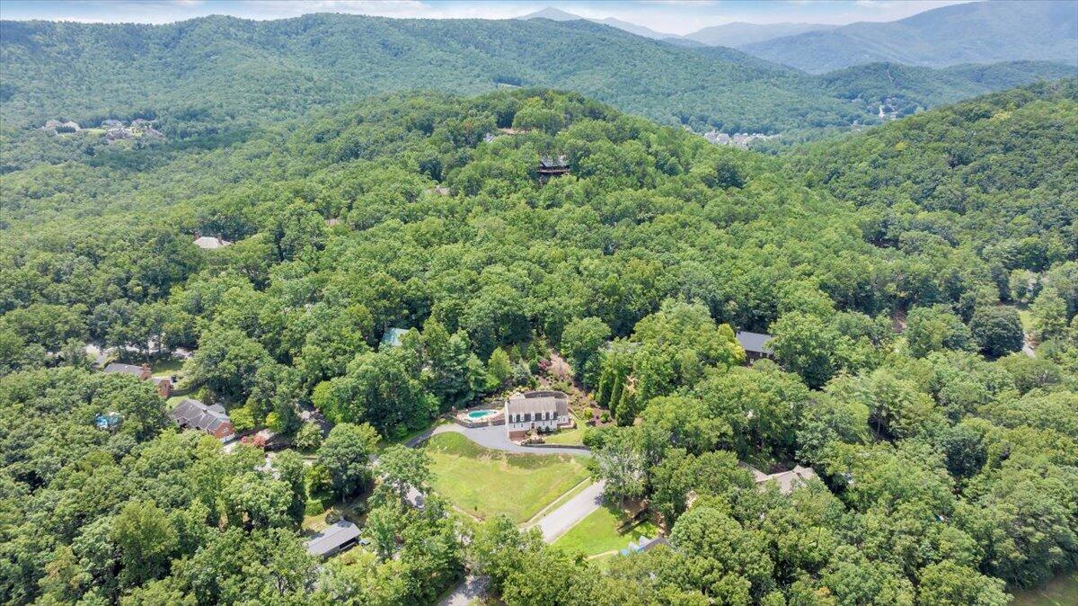 Aerial view of a home amid dense woodland, with a pool and mountain ridges beyond.