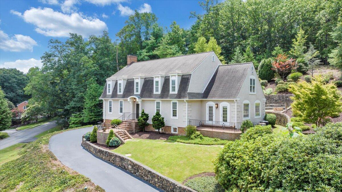 Light-colored house with roof dormers, a curved driveway, and a stone retaining wall amid mature trees.
