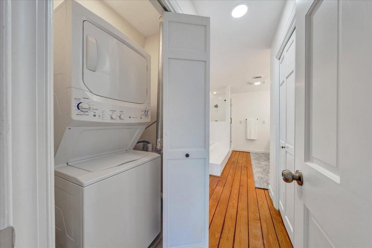 Stacked washer and dryer beside a bathroom, with warm wood plank flooring.