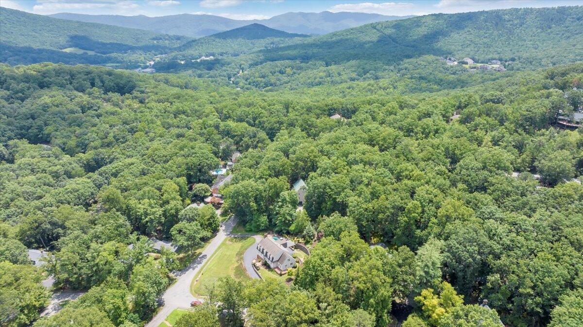 Aerial view of a house amid dense trees, with forested mountain ridges in the distance.