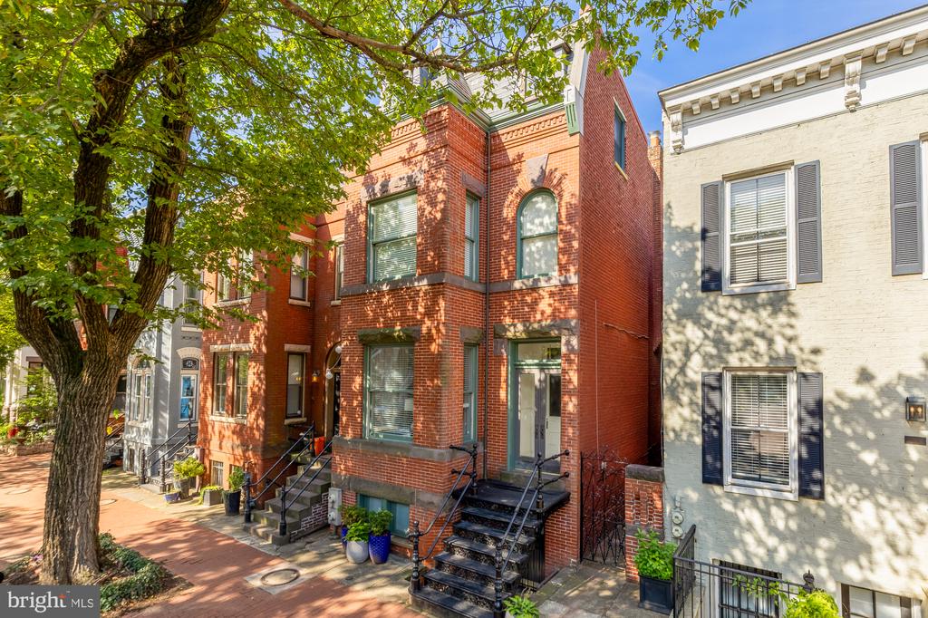 Red-brick townhouse with a raised entry and black railings beneath a leafy street tree.