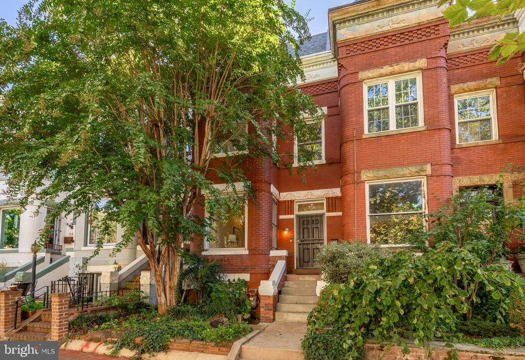 Red brick rowhouse with a projecting bay, stone-trimmed windows, front steps, and mature greenery.