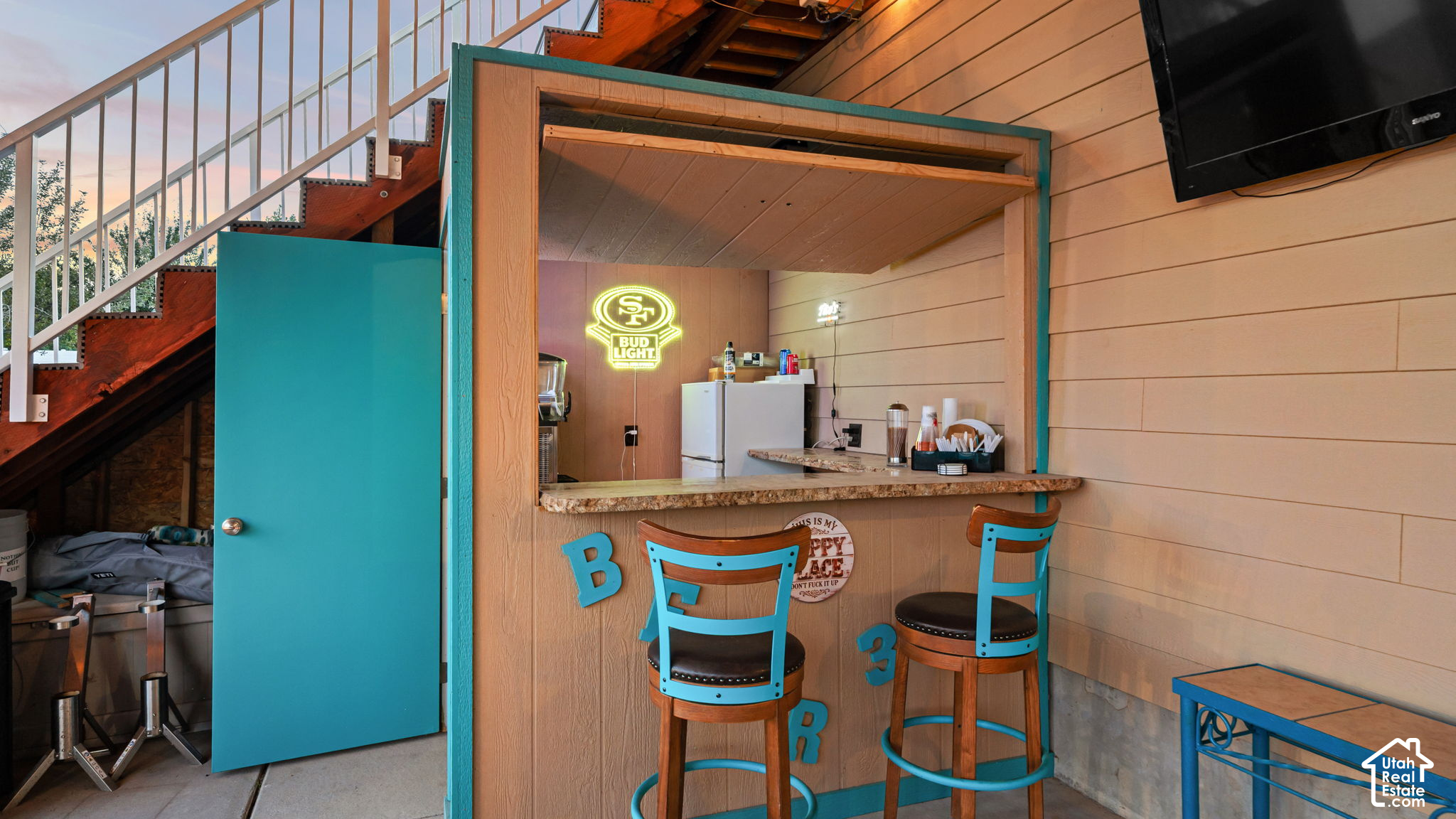 Covered outdoor bar beneath a staircase, with turquoise stools and a glowing 49ers Bud Light sign.