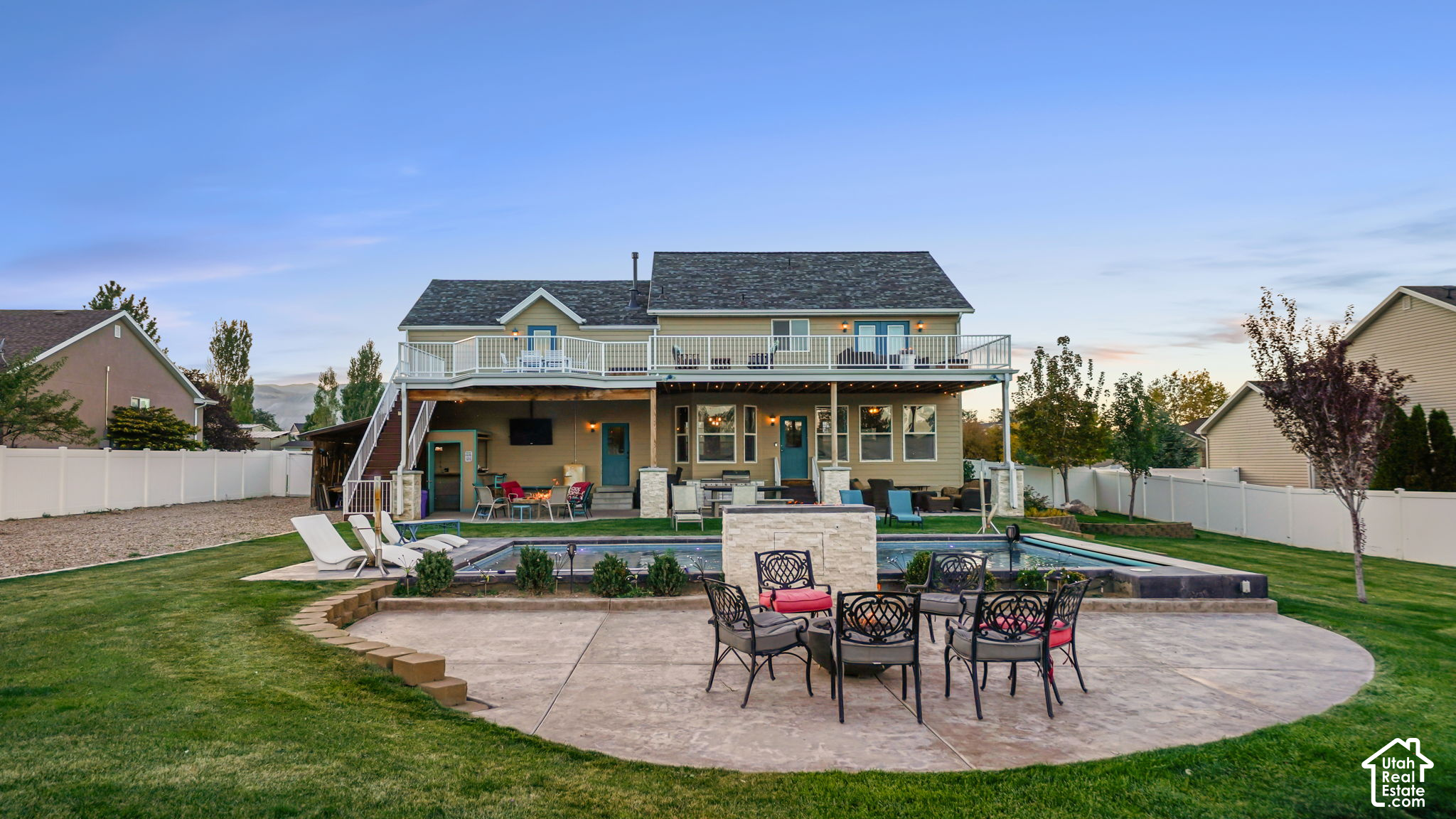 Home’s rear exterior with an elevated deck, fenced pool, and patio seating at dusk.