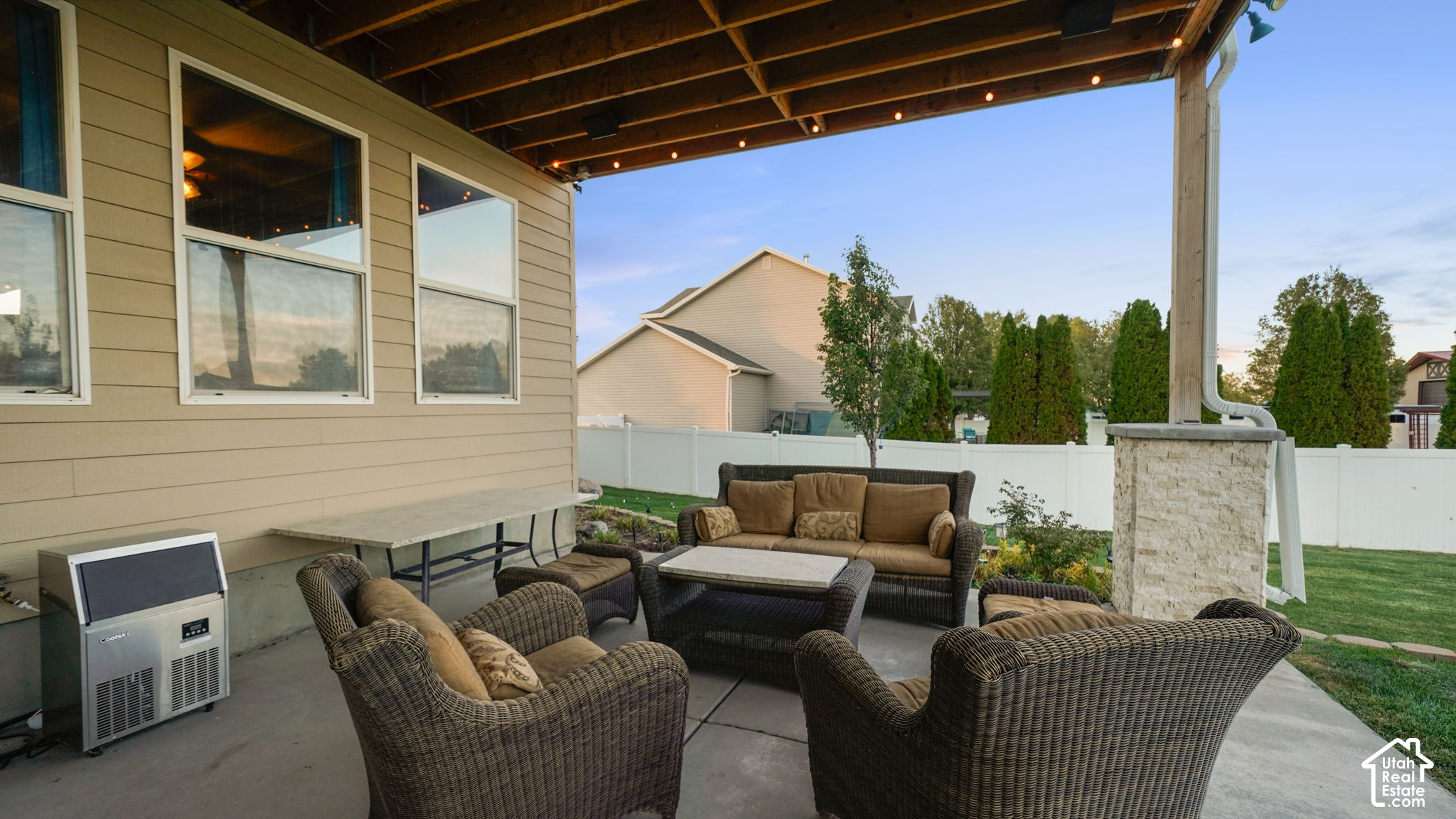 Covered patio with wicker seating, string lights, and a stone-clad column beside a fenced yard.